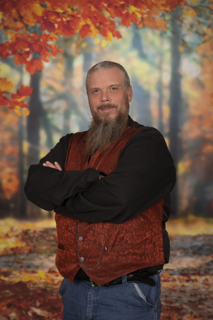 A man with a long beard, wearing a patterned red vest and black shirt, stands with arms crossed in front of an autumn forest backdrop.