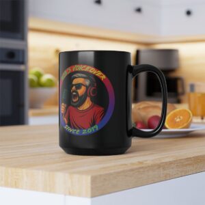 A black mug on a kitchen counter features an illustration of a man with headphones and a microphone, with the text “Rob’s Voiceover Since 2019.” Fruit and appliances are in the background.