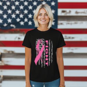 A woman stands before a large American flag, wearing the Pink Ribbon Fight T-Shirt—a black tee featuring a pink ribbon and the word "FIGHT.
