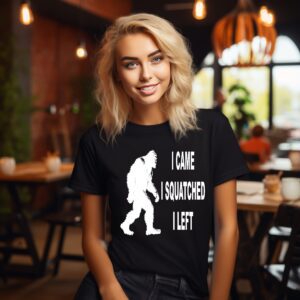 A woman sits in a cafe wearing the Squatch Tee, a black T-shirt featuring a white silhouette of Bigfoot and the text "I CAME I SQUATCHED I LEFT.