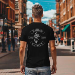 A man faces away on a city street, wearing the Wisconsin Stagehand Local Crew Tee—a black shirt featuring "Stagehand Local Crew Wisconsin" and a deer head graphic.