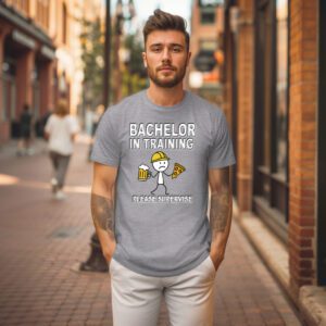 A man stands on a city sidewalk wearing the Bachelor in Training T-Shirt, which features the phrase "Bachelor in Training, Please Supervise" along with cartoon images of a person, pizza, and a beer mug.