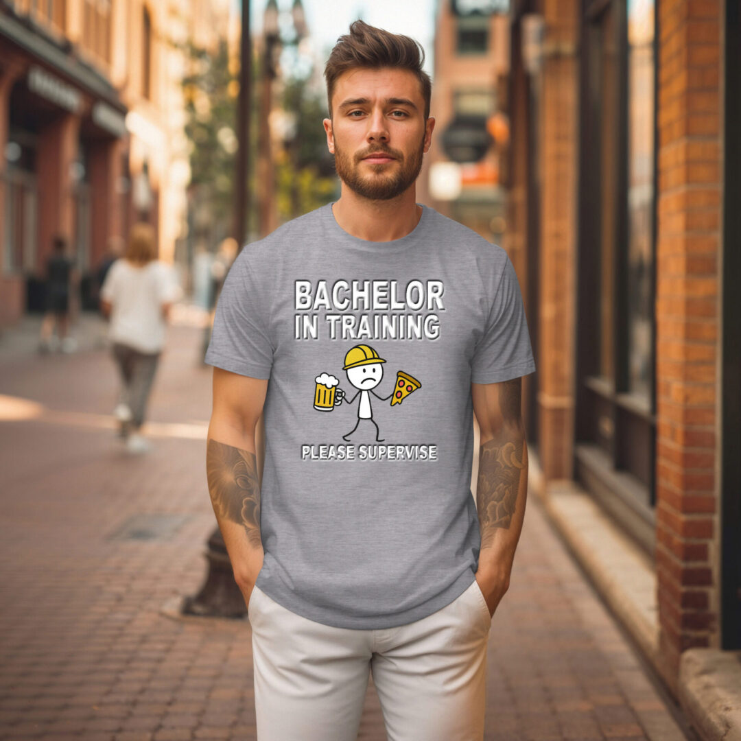 A man stands on a city sidewalk wearing the Bachelor in Training T-Shirt, which features the phrase "Bachelor in Training, Please Supervise" along with cartoon images of a person, pizza, and a beer mug.