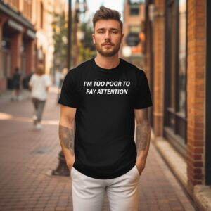 A man stands on a brick-paved city street wearing the Too Poor Tee, a black t-shirt with the phrase "I'M TOO POOR TO PAY ATTENTION" printed in white letters.