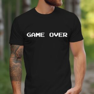 A bearded, tattooed man stands outdoors on a wooded path wearing the Game Over Tee, a black T-shirt with "GAME OVER" in white pixelated font.