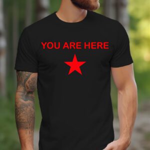 A tattooed man wearing the Guiding Star Tee—a black shirt with "YOU ARE HERE" above a red star—stands outdoors on a woodland path.