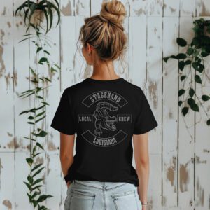 A woman faces away from the camera, wearing the Louisiana Stagehand Local Crew Tee featuring an alligator graphic, standing before a white wooden wall with green plants.