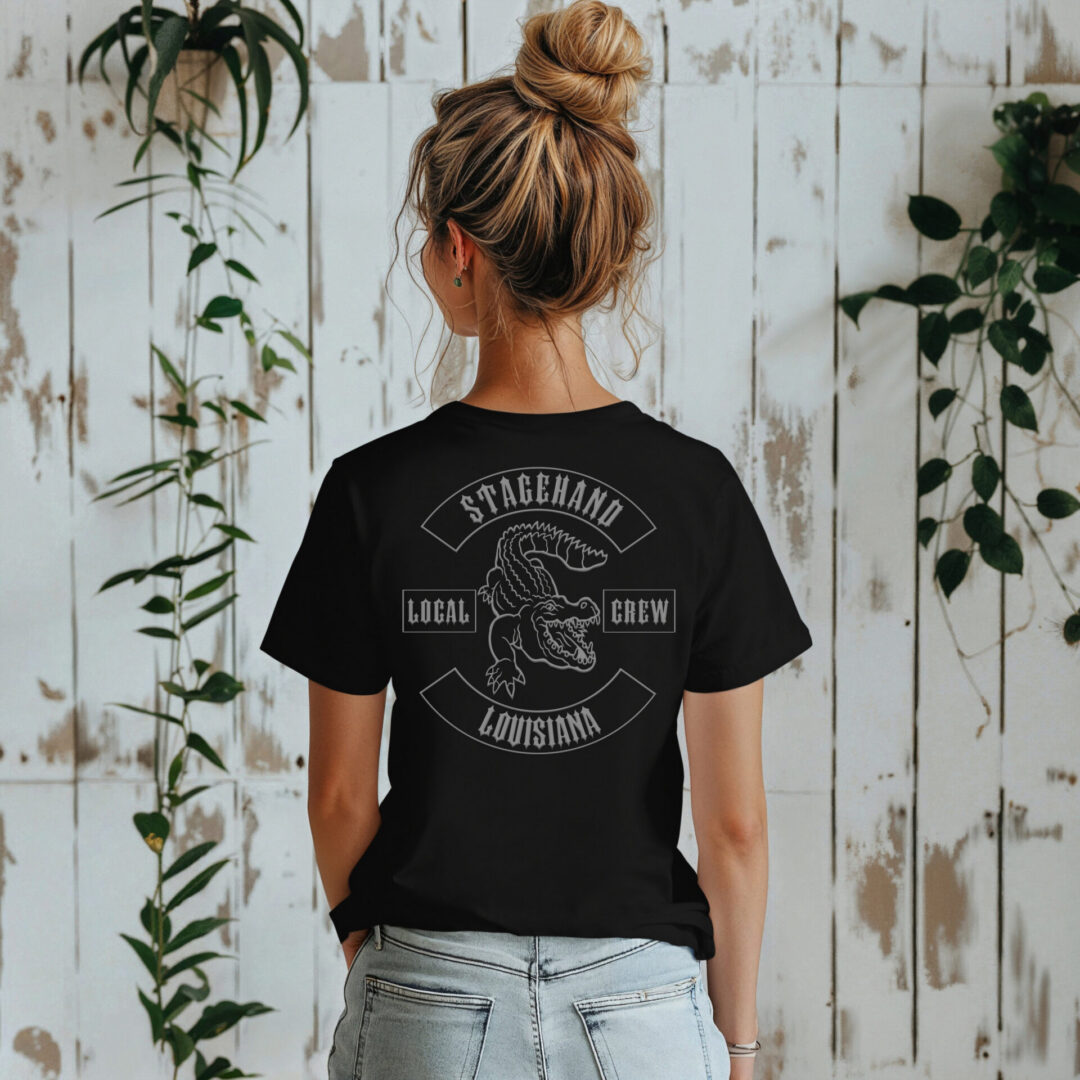 A woman faces away from the camera, wearing the Louisiana Stagehand Local Crew Tee featuring an alligator graphic, standing before a white wooden wall with green plants.