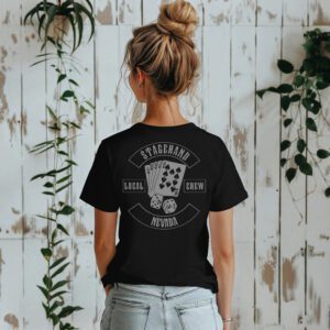 A woman stands indoors facing a white wooden wall with green vines, wearing the Nevada Stagehand Local Crew Tee featuring playing cards and dice graphics on the back.