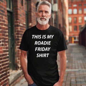 A man with gray hair and a beard stands in an alley wearing the "This Is My Roadie Friday Tee," a black t-shirt with the phrase printed in white letters.