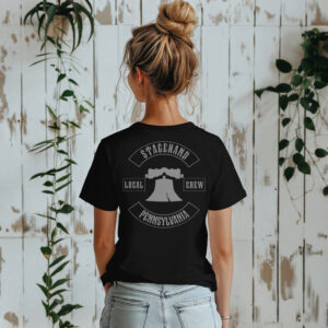 A woman with a messy bun faces a white wooden wall, wearing the Pennsylvania Stagehand Local Crew Tee and light blue jeans.