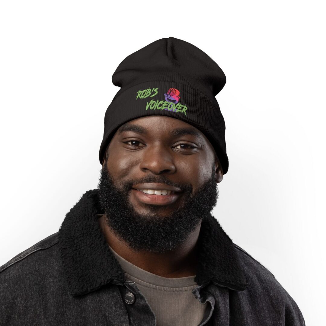 A bearded man smiles at the camera against a white background, wearing the Rob's Voiceover Embroidered Knit Beanie featuring "Rob's Voiceover" text and a pink microphone graphic.